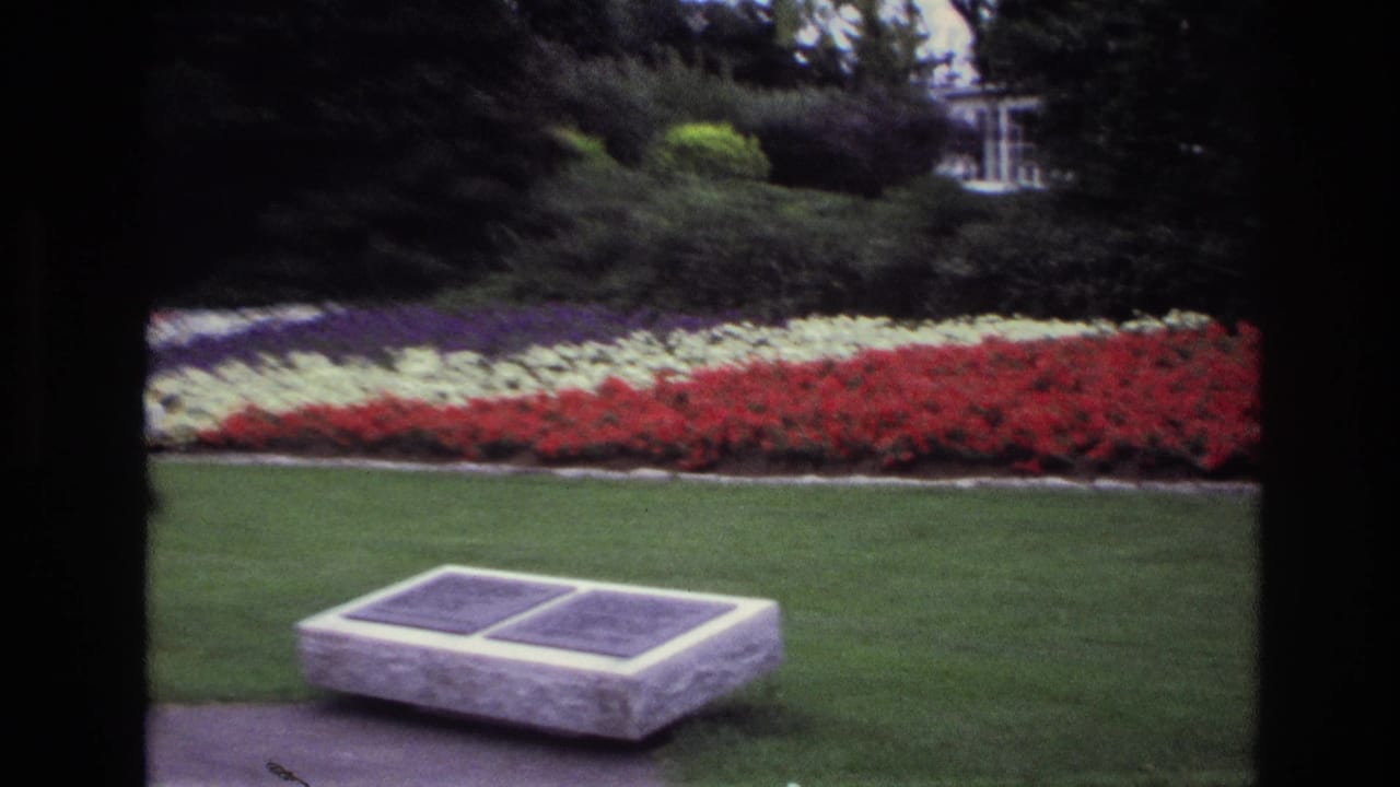 1967 Ottawa garden view with a stone memorial bench, two plaques, clipped lawn, layered flowerbeds, shrubs, and part of a building beyond.