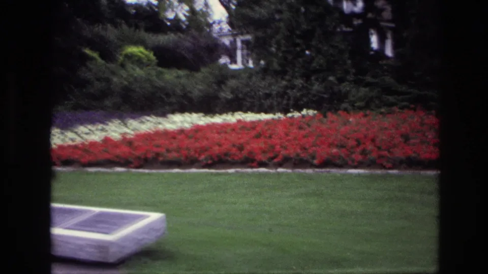 1967 Ottawa 8mm frame showing the edge of a memorial bench in the foreground, broad lawn, bright flowerbeds, shrubs, and building details behind.