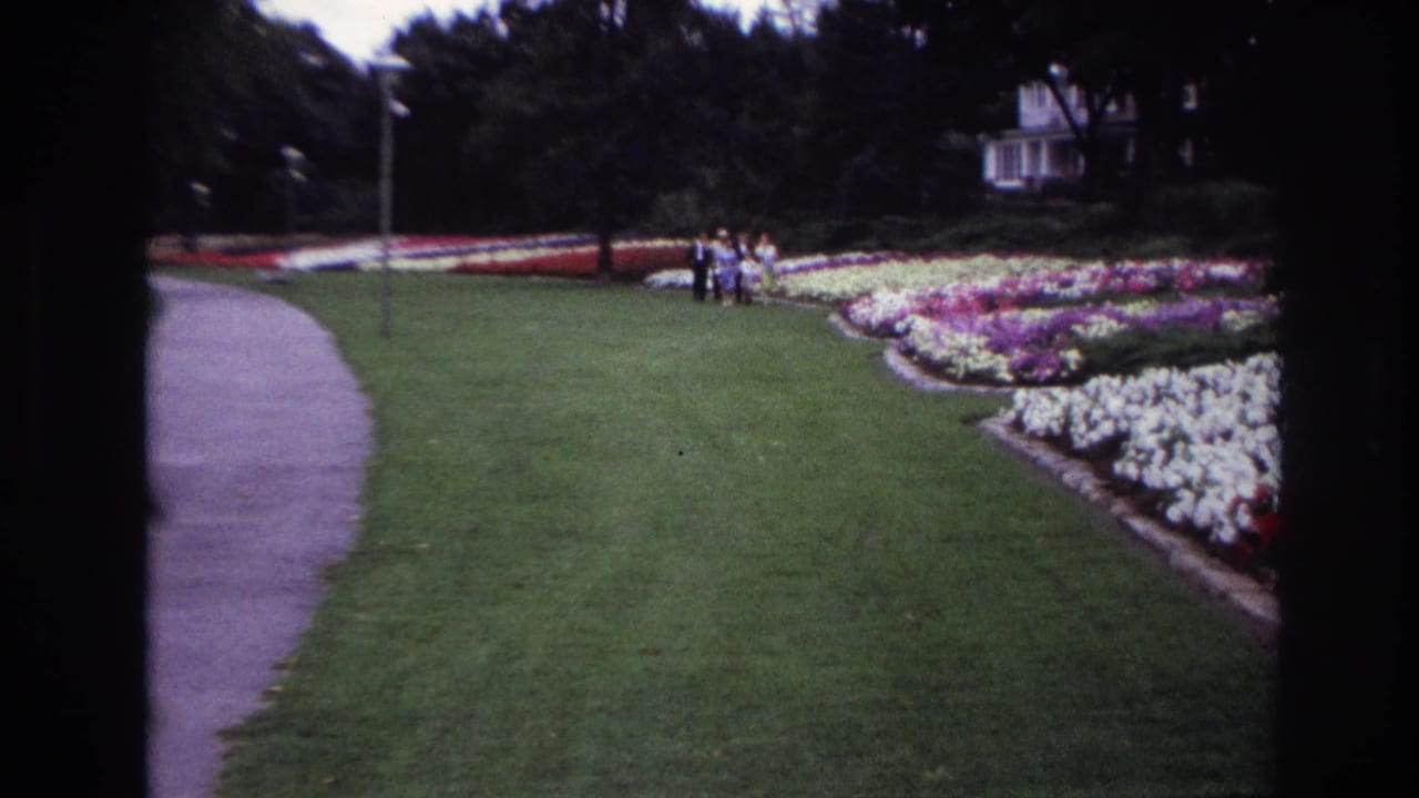 Ottawa 1967 home-movie frame showing a small distant group near colorful garden beds, a curved path, lawn, trees, and a white house-like building.