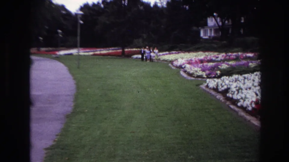 Ottawa 1967 home-movie frame showing a small distant group near colorful garden beds, a curved path, lawn, trees, and a white house-like building.