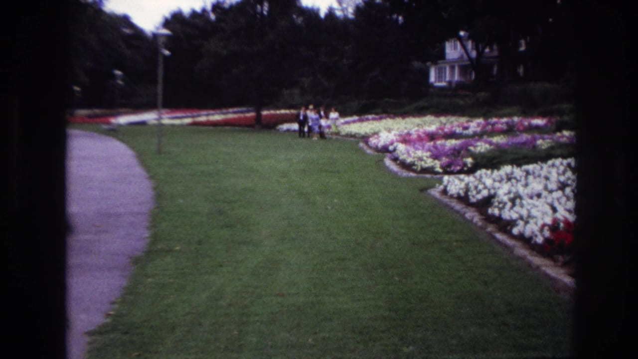Ottawa 1967 frame with distant visitors among patterned flower beds, bordered by a curved paved path and mature trees.