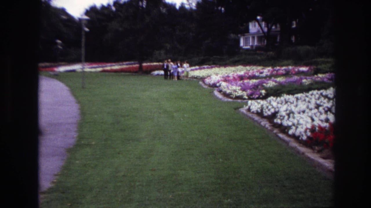Ottawa 1967 garden frame showing distant people near sweeping flower beds, a broad lawn, curved path, trees, and a white house-like building.