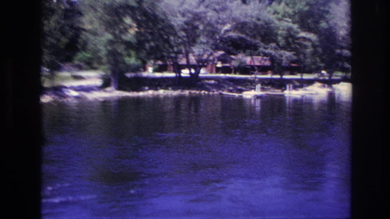 Ottawa-area 1967 home-movie frame showing blue lake water, trees, and small lakeside buildings along the opposite shore.