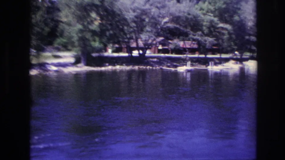 Ottawa-area 1967 home-movie frame showing blue lake water, trees, and small lakeside buildings along the opposite shore.