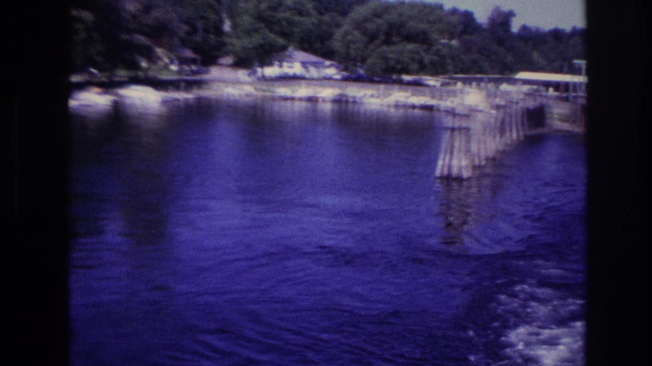 Ottawa 1967 8mm frame showing a small dock or pier extending into blue lake water beside a tree-lined shore.