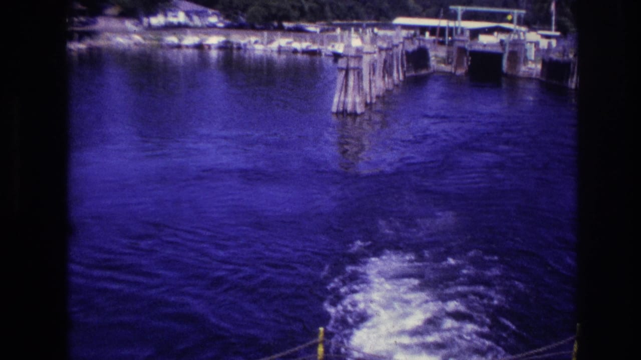 1967 Ottawa home-movie frame with wooden dock posts, shoreline structures, and boat wake across blue-tinted lake water.
