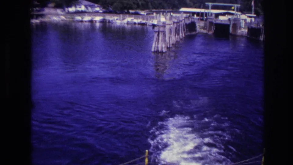 1967 Ottawa home-movie frame with wooden dock posts, shoreline structures, and boat wake across blue-tinted lake water.