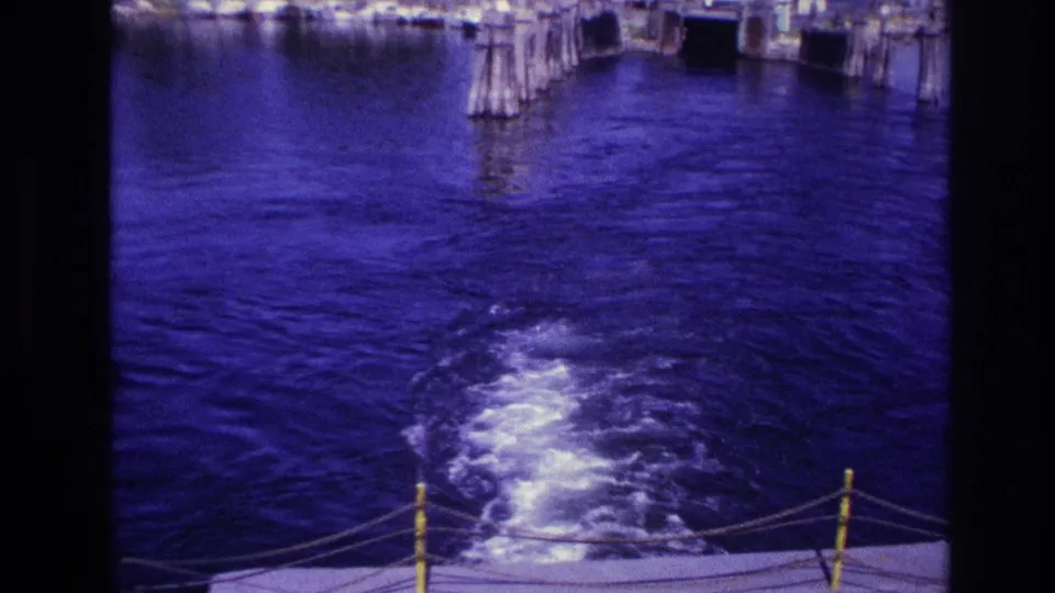 1967 Ottawa-area home-movie frame showing a boat wake churning behind the stern as wooden dock pilings recede.