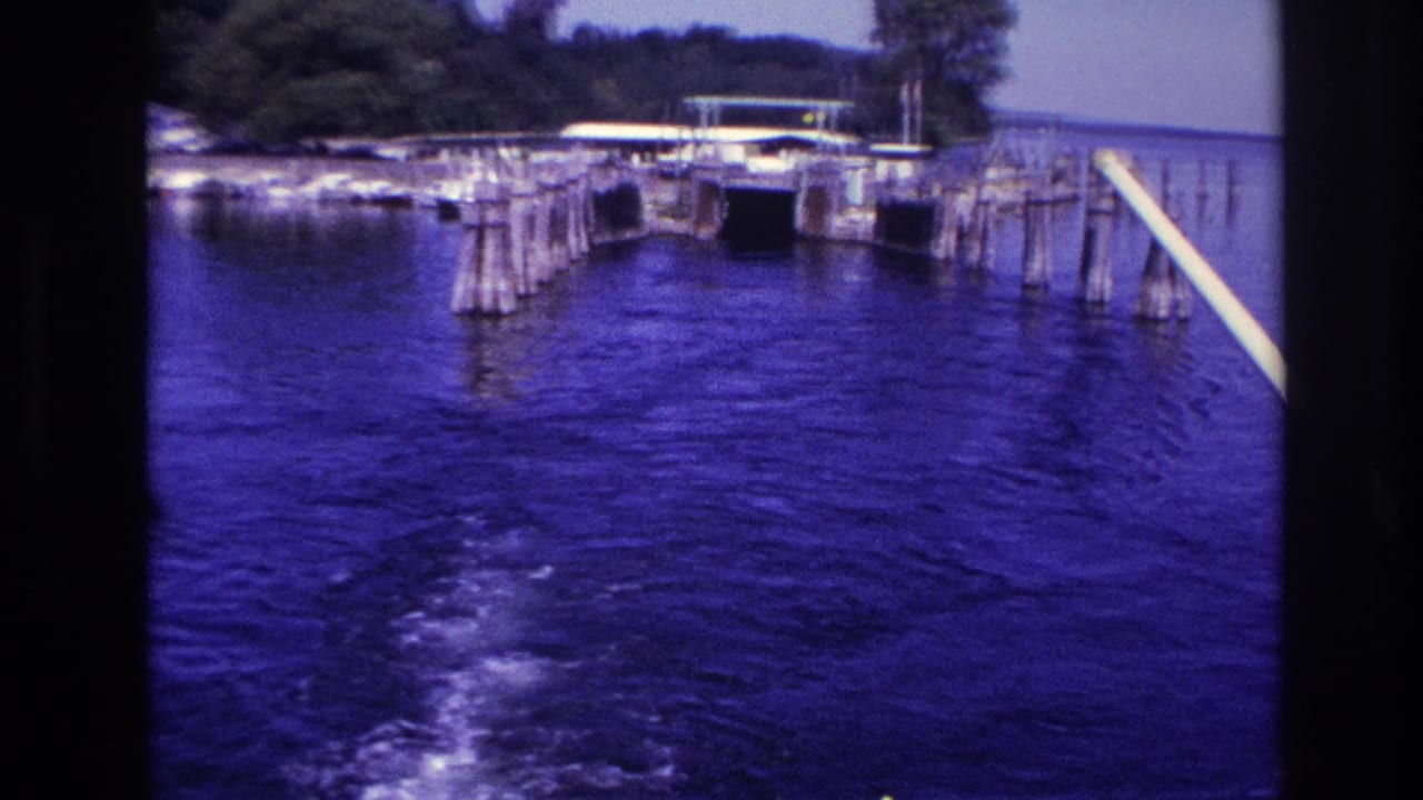 1967 Ottawa-area boating view with the wake fading across blue water near wooden piers and a small shoreline structure.