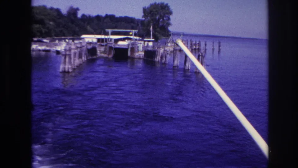 1967 Ottawa-area home-movie frame showing a pale boat rail, dock pilings, shoreline trees, and open water.