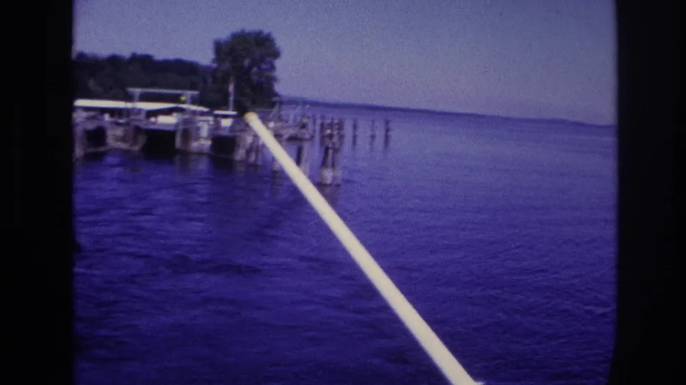 1967 Ottawa-area frame with open blue water, a diagonal boat rail, distant dock pilings, and a wooded shoreline.