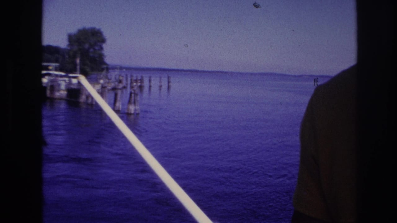 1967 Ottawa-area 8mm frame looking from a boat across calm water toward dock pilings and a wooded shore, with a white rail crossing the foreground.