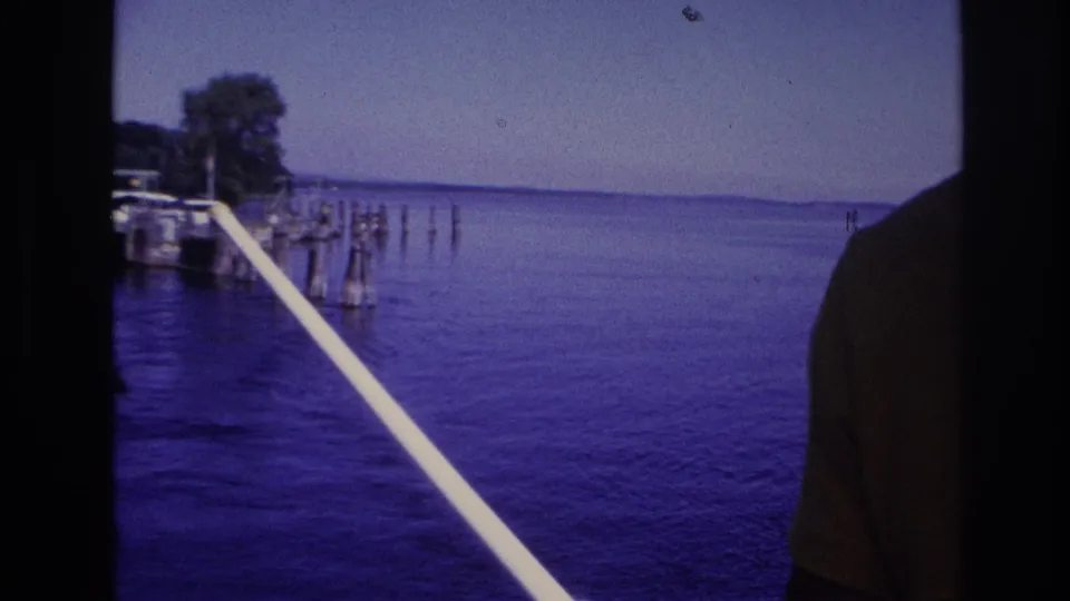 1967 Ottawa-area 8mm frame looking from a boat across calm water toward dock pilings and a wooded shore, with a white rail crossing the foreground.
