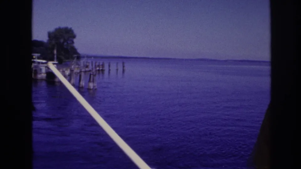 1967 Ottawa-area home-movie frame showing open blue water, a white rail, dock pilings, and distant wooded shoreline from a boat perspective.