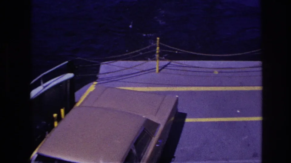 1967 Ottawa-area ferry deck seen from above, with a car roof, yellow painted lines, rope railings, and dark water behind.