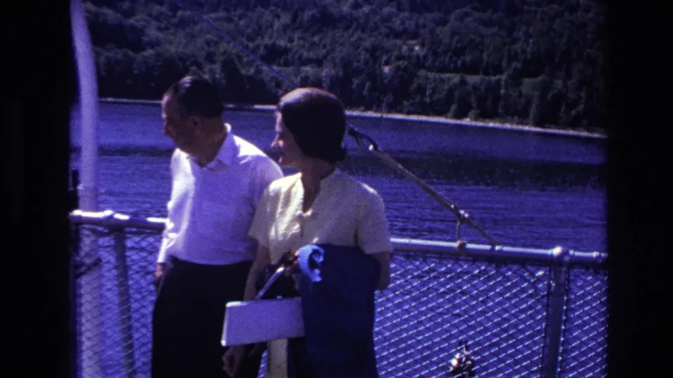1967 Ottawa home-movie frame showing the couple by a ferry railing, with water, trees, and deck hardware behind them.