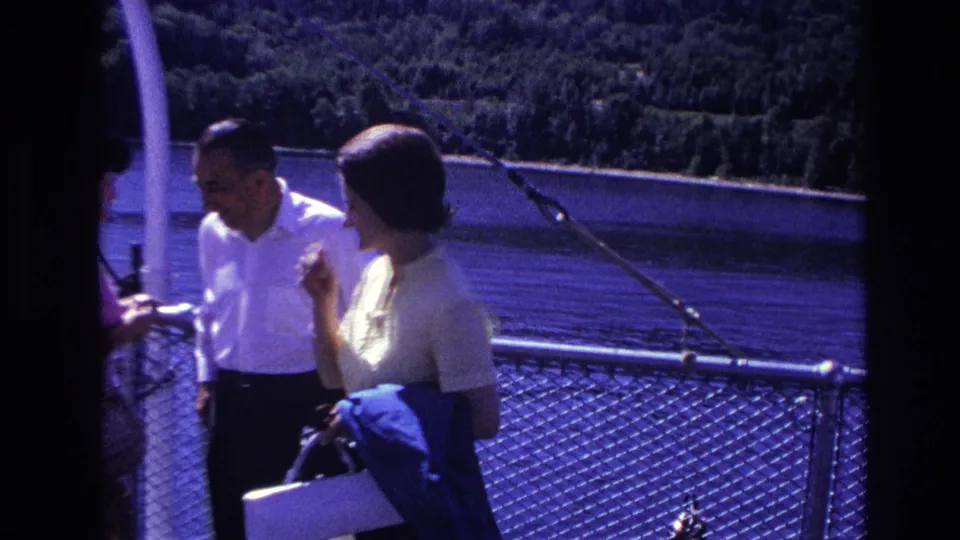 In Ottawa, Canada, in 1967, a man in a white shirt and a woman in a light dress stand near a boat railing by calm water.