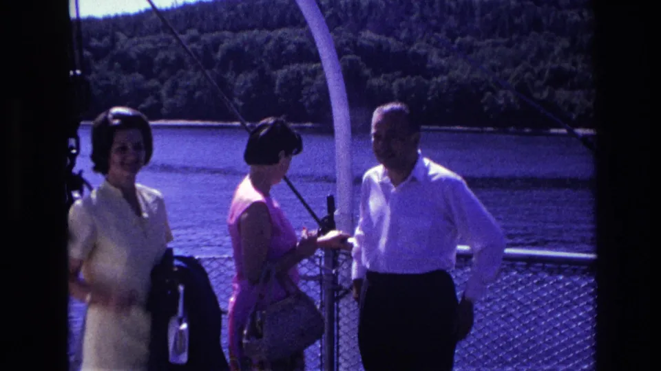 Three adults aboard a boat in 1967 Ottawa footage stand near the railing with water and wooded shoreline in the background.