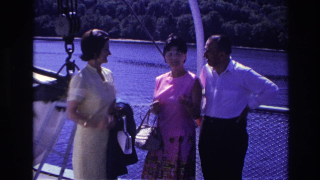 Three adults stand on a boat deck beside open water and a wooded shoreline.