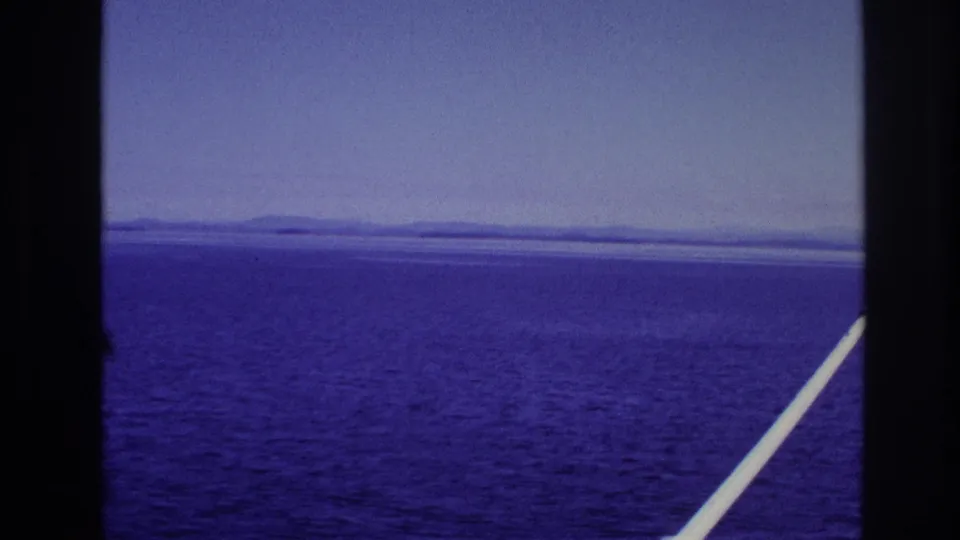 1967 8mm travel view of blue-purple water and a distant shoreline framed by dark window bars and a white diagonal pole.