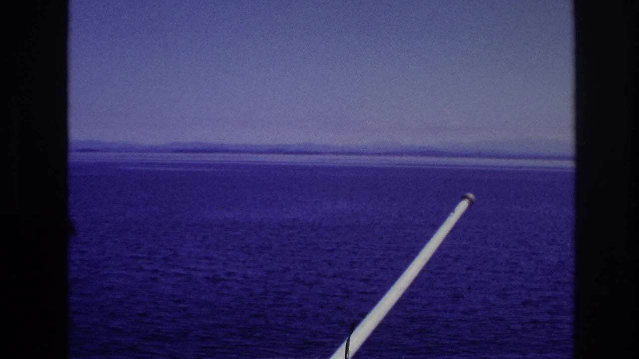 Color-shifted 1967 8mm frame showing water, horizon, and distant land through dark window dividers with a white pole in the foreground.