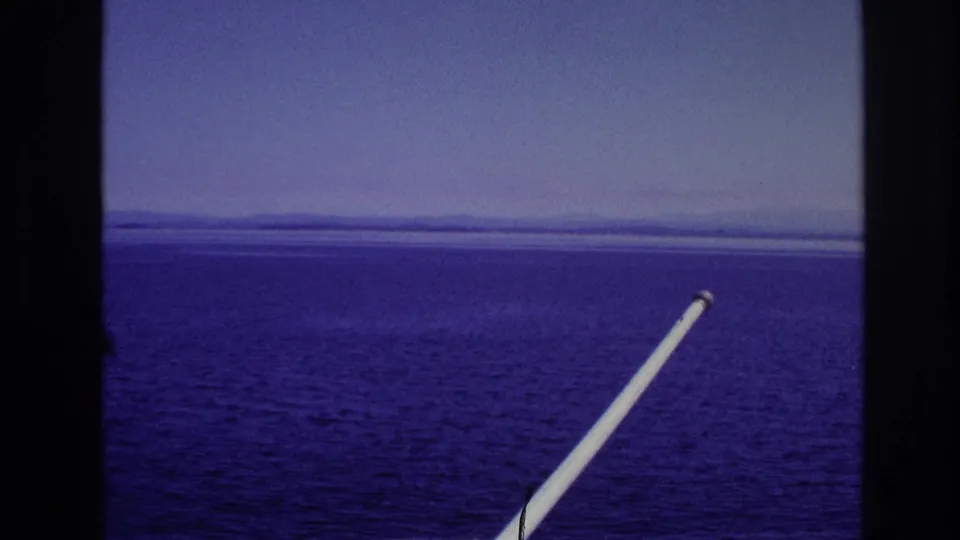 Color-shifted 1967 8mm frame showing water, horizon, and distant land through dark window dividers with a white pole in the foreground.