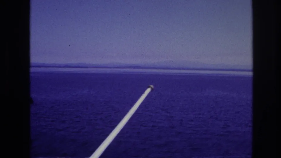 1967 home-movie frame of open water and distant shoreline, with dark framing edges and a white diagonal railing in front.