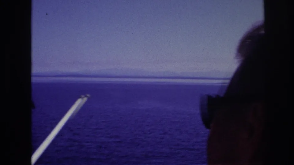 1967 home-movie frame showing passengers in silhouette looking over open water from a moving boat, with a white pole at left.