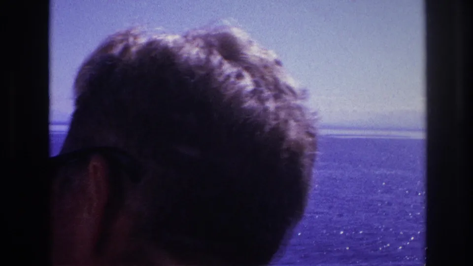 A man with short hair and sunglasses is seen in close silhouette while looking toward open water in 1967 travel footage.