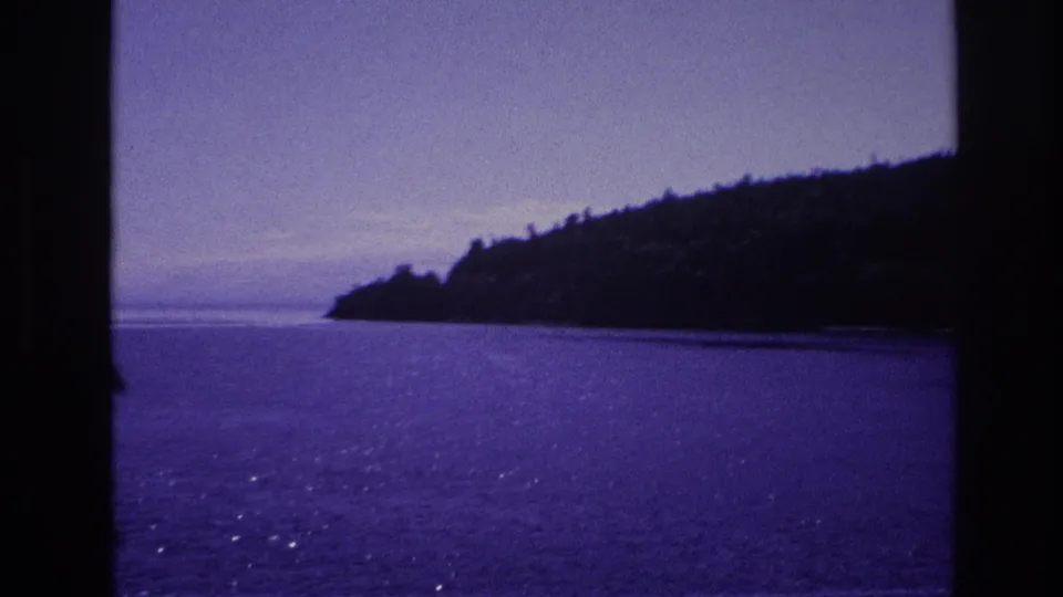 A purple-blue 8mm frame shows open water and a low wooded shoreline viewed from a boat, with no clear person in this frame.