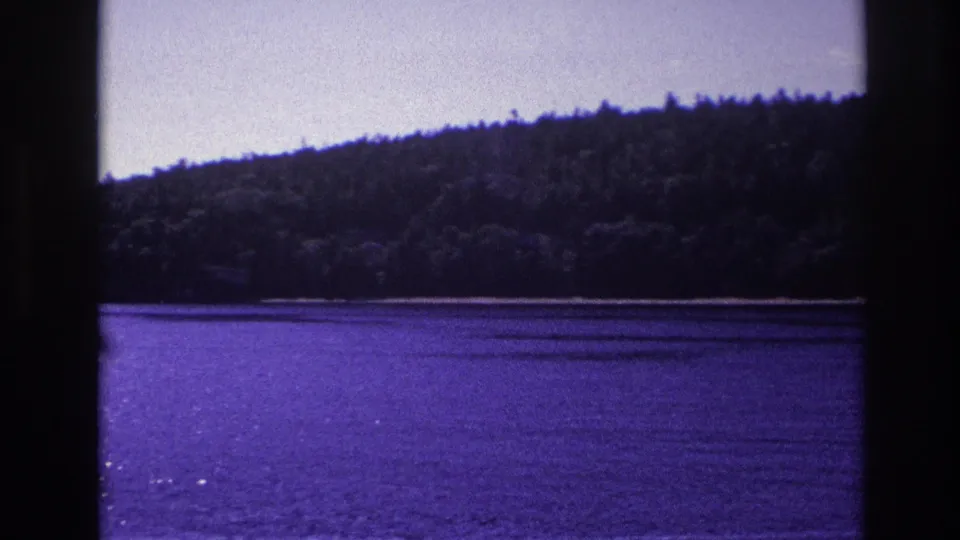 1967 Ottawa home-movie still with a wooded lakeshore across purple-blue water, partly blocked by dark window framing.
