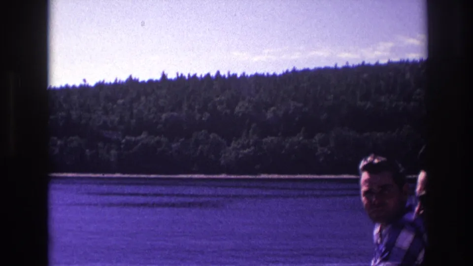 1967 Ottawa home-movie frame showing a man on a boat with a lake and tree-covered hillside in the background.