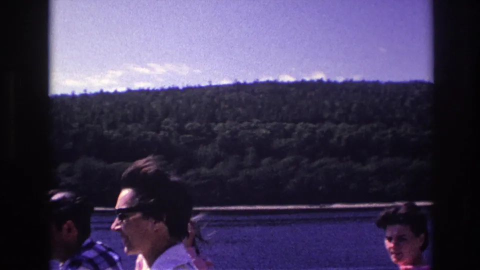 1967 Ottawa home-movie view of boat passengers, including a woman in sunglasses, with lake and hillside beyond.