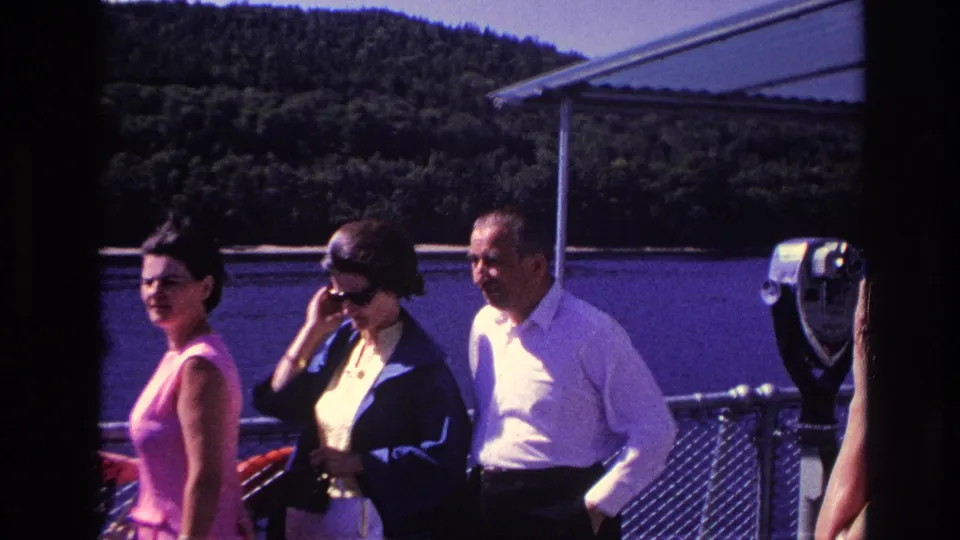 1967 Ottawa home-movie frame showing three adults near a boat railing, with river water and a wooded hillside in the background.