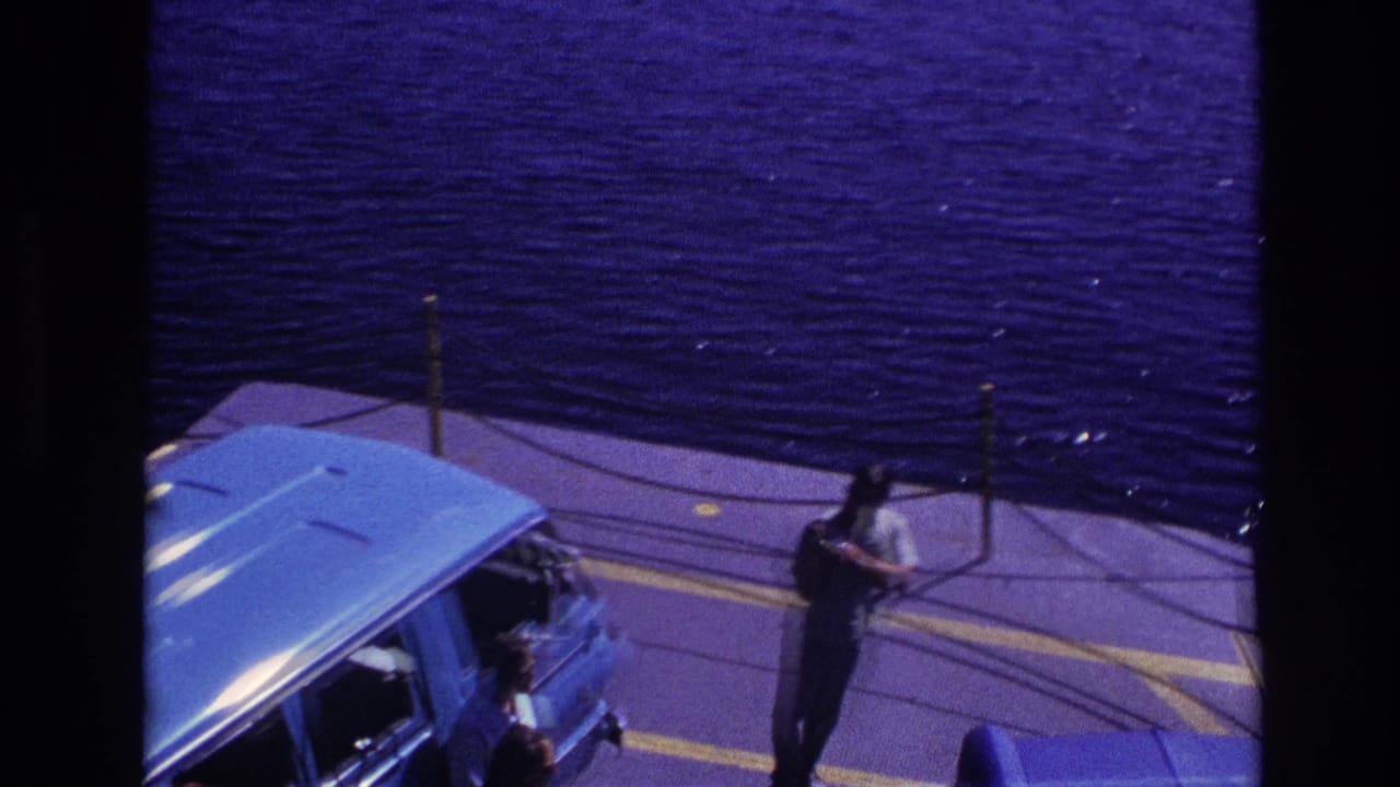 1967 Ottawa home-movie frame showing a man holding a bag near a blue car on a ferry deck with water behind him.