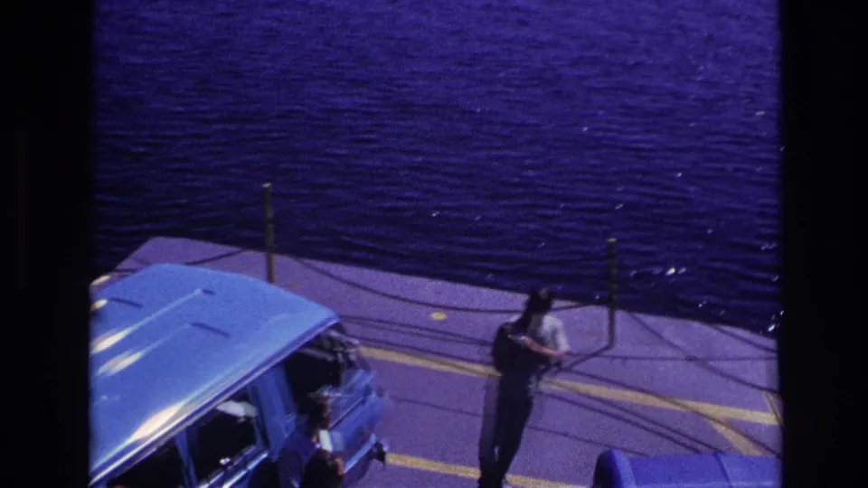 1967 Ottawa home-movie frame showing a man holding a bag near a blue car on a ferry deck with water behind him.