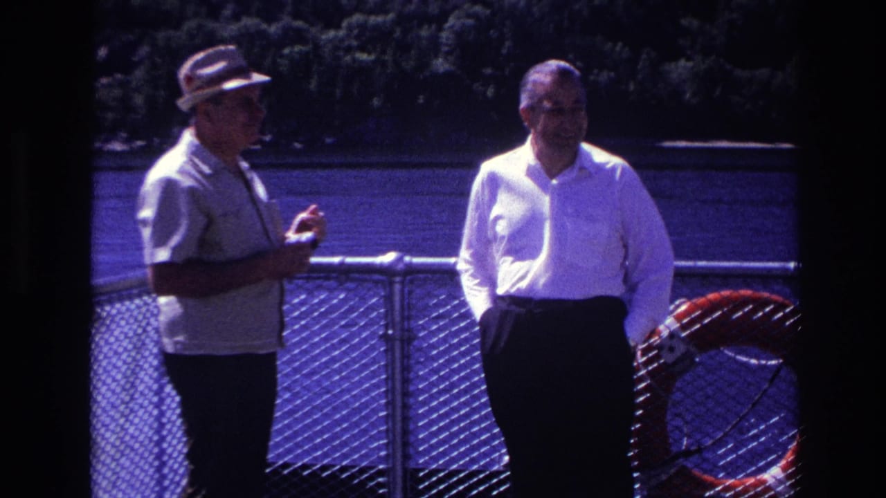 Two men continue talking beside a railing with water behind them.