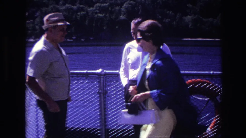 A woman in sunglasses and a dark jacket enters the frame near two men by the water in Ottawa, Canada, 1967.