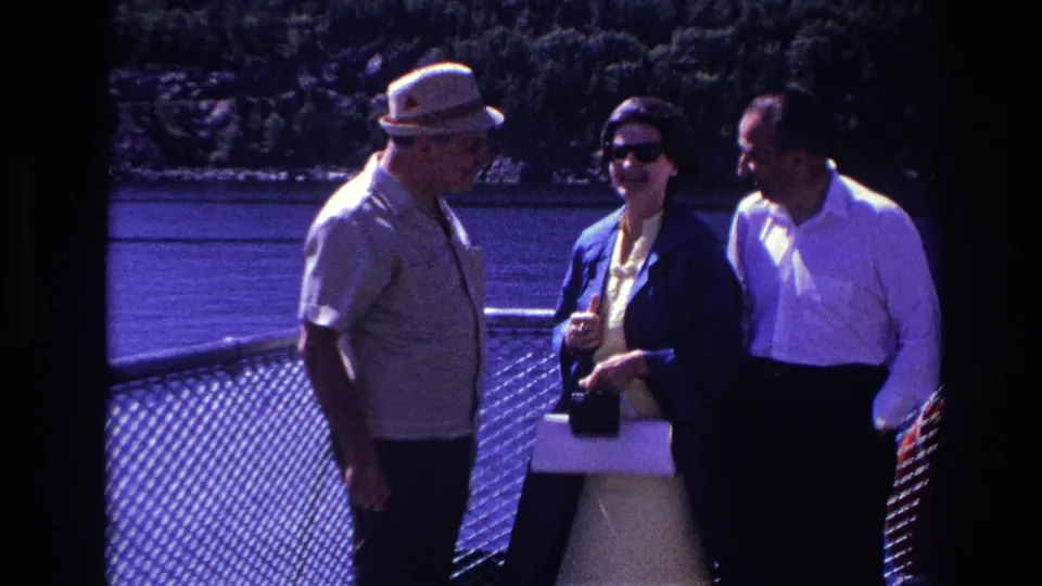 Three adults in 1967 Ottawa footage smile and talk on a boat deck, the woman wearing dark sunglasses and holding a purse.