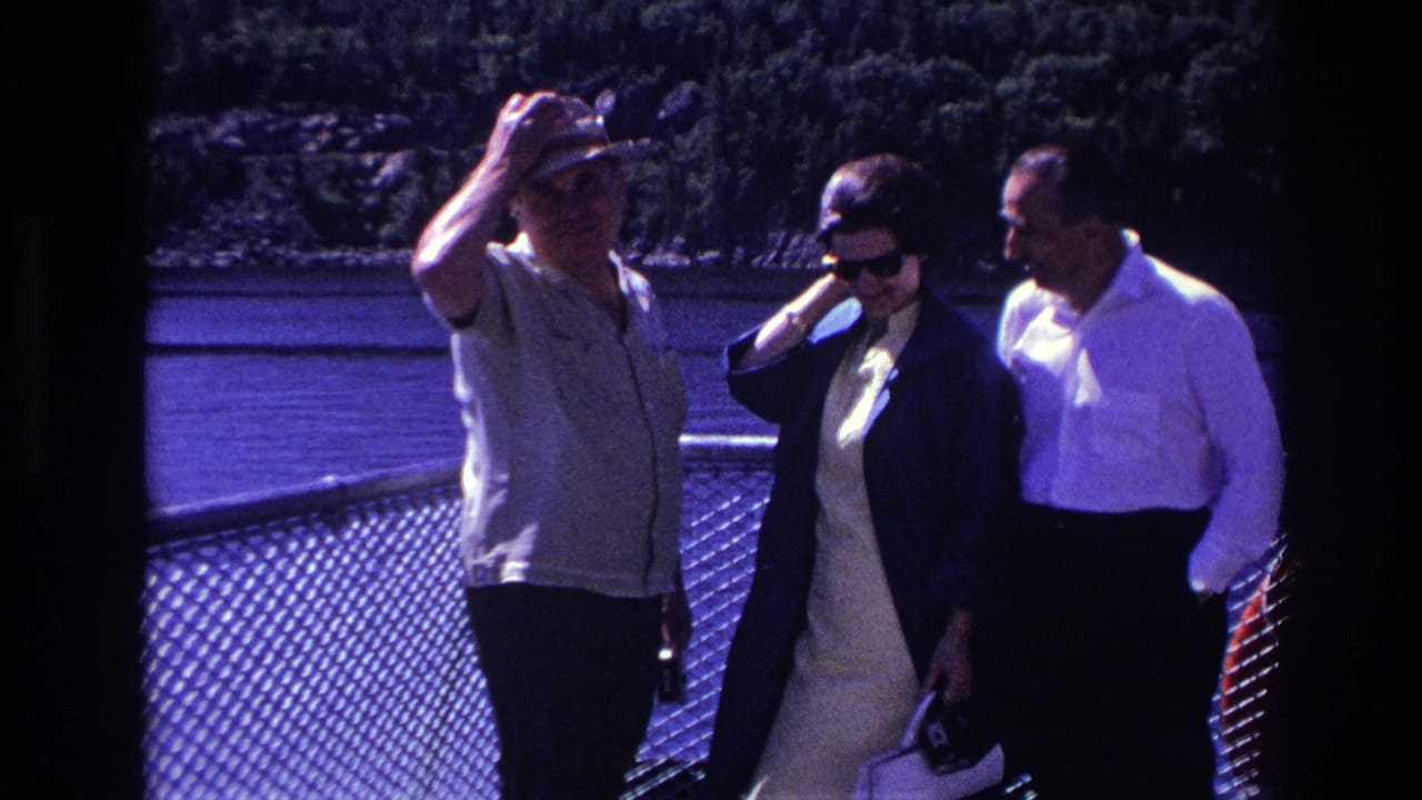 1967 home-movie frame of adults on a boat deck, including a man shielding his eyes and two passengers in sunglasses by the railing.