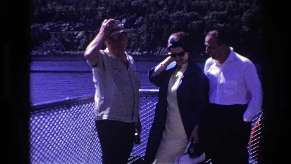 1967 home-movie frame of adults on a boat deck, including a man shielding his eyes and two passengers in sunglasses by the railing.