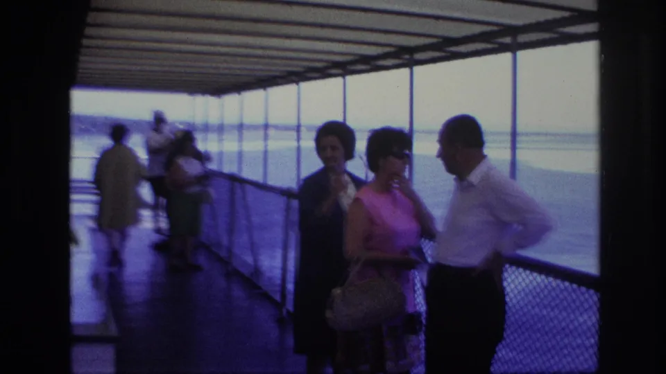 Home-movie frame of adults gathered along a covered boat deck, with a woman in a pink dress holding a handbag near the railing.