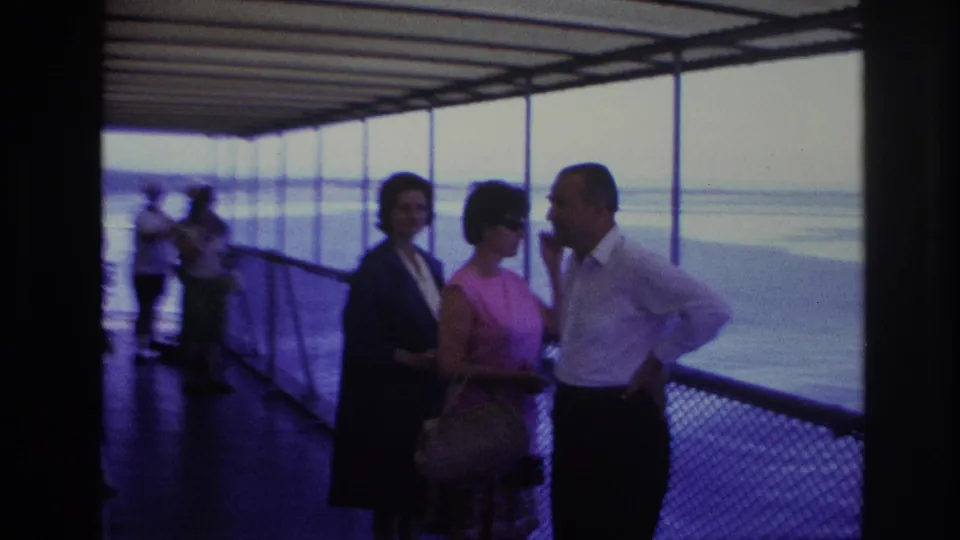 In Ottawa, Canada in 1967, a man in a white shirt and two women stand talking on a covered boat deck by large windows.