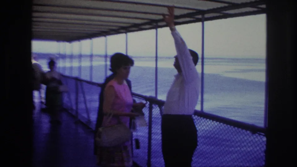 During the Ottawa boat ride, the man raises his arm toward the view while the woman in pink stands nearby with a handbag.