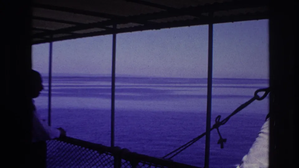 Underexposed 8mm frame shows visitors in silhouette at the covered railing, with blue water and a hazy horizon beyond.