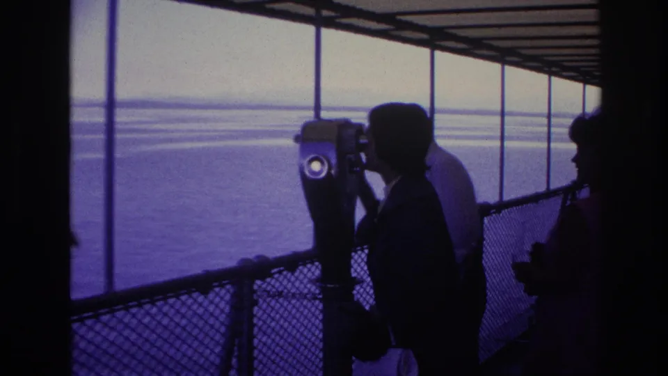 Blue-purple 8mm frame of a tourist looking through a fixed pier viewer with companions silhouetted against the water.