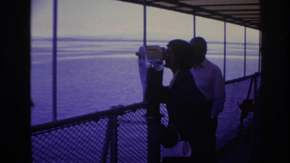 1967 waterfront travel frame showing a visitor at a fixed binocular viewer under a covered pier, with water beyond.