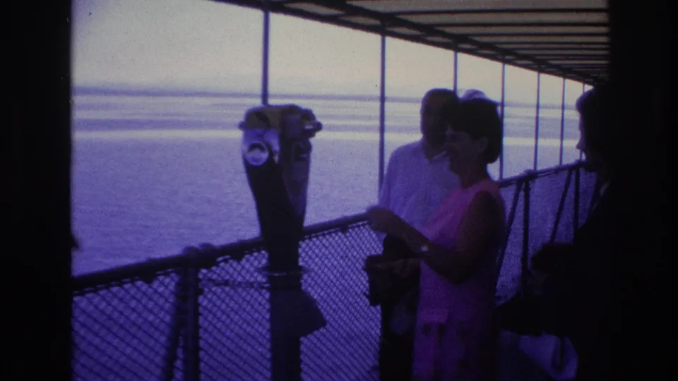 1967 Ottawa home-movie frame showing adults on a covered deck as a woman in pink stands near a binocular viewer overlooking water.