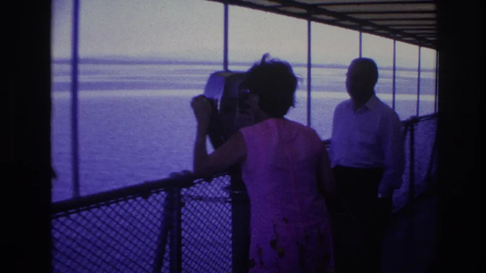1967 Ottawa home-movie frame showing the woman from behind at the binocular viewer, with a man standing near the rail and water beyond.
