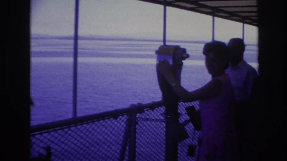 The woman in the pink dress adjusts the mounted viewer on the boat deck, with another adult partly visible and open water beyond the railing.
